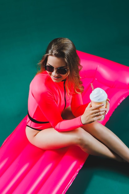 Mulher muito magra em um maiô rosa encontra-se em um colchão inflável com um coquetel nas mãos em um fundo verde isolado Férias de verão no conceito de bronzeamento do mar Colchão de ar rosa