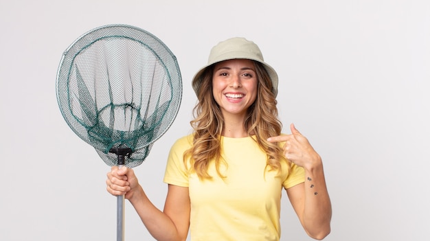 Mulher muito magra se sentindo feliz e apontando para si mesma com um chapéu animado e segurando uma rede de pesca