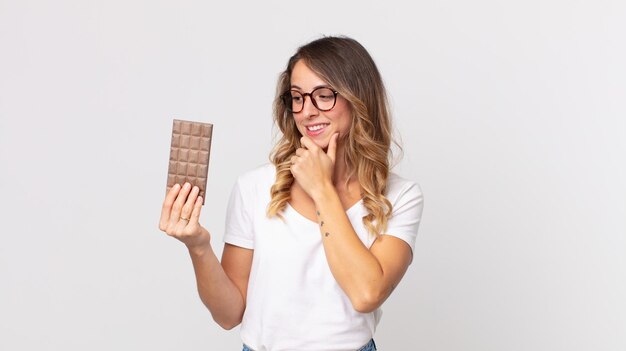 Mulher muito magra sorrindo com uma expressão feliz e confiante com a mão no queixo e segurando uma barra de chocolate