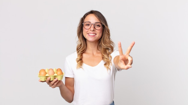 Mulher muito magra sorrindo e parecendo feliz, gesticulando vitória ou paz e segurando uma caixa de ovos