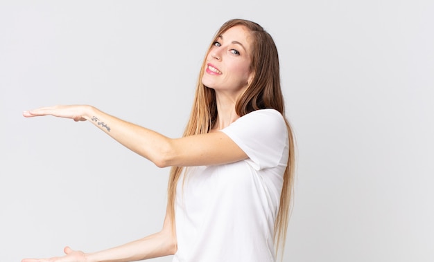 Mulher muito magra sorrindo, sentindo-se feliz, positiva e satisfeita, segurando ou mostrando um objeto ou conceito no espaço da cópia