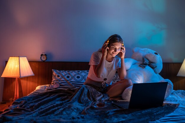 Mulher muito sorridente ocupado navegando no laptop com fones de ouvido enquanto toma uma xícara de chá na cama aconchegante.