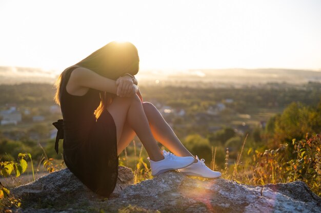 Mulher muito triste com vestido preto curto de verão, sentado em uma pedra, pensando ao ar livre ao pôr do sol. Mulher na moda, contemplando a noite quente na natureza.