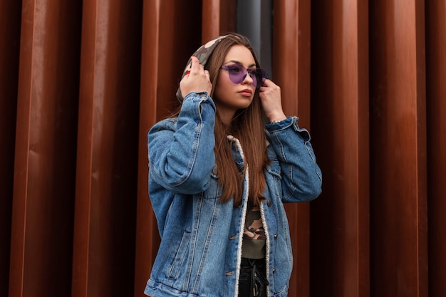 Mulher muito urbana jovem hippie coloca um capuz. Modelo de menina bonita com elegante jaqueta jeans com glamourosos óculos roxos se passando perto de uma moderna parede de metal ao ar livre. Estilo de rua moderno da juventude casual.