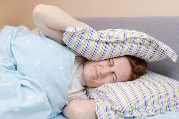 Mulher na cama cobriu a cabeça com travesseiro e fechou os olhos. O barulho dos vizinhos impede a mulher de dormir. Má insonorização em edifícios de apartamentos. Mulher se irrita com barulho e não consegue dormir