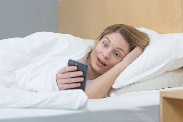 Mulher na cama debaixo do cobertor chocada lendo boas notícias do telefone sorrindo