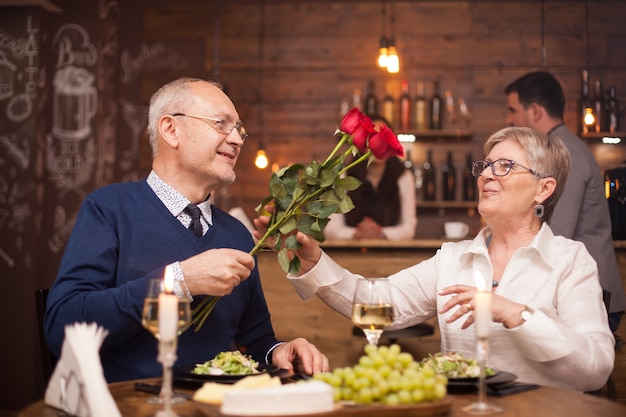 Mulher na casa dos sessenta anos feliz por receber rosas do marido durante o jantar. encontro do casal sênior. uvas frescas. casal idoso alegre.