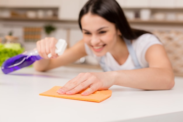 Mulher na cozinha está sorrindo e limpando a poeira com um spray e um espanador enquanto limpa a casa, close-up