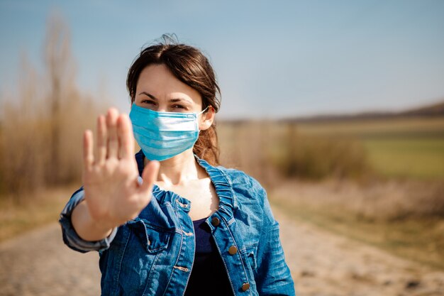 Mulher na máscara médica estéril protetora no rosto, olhando para a câmera ao ar livre. Parada de mão Conceito de coronavírus pandêmico.