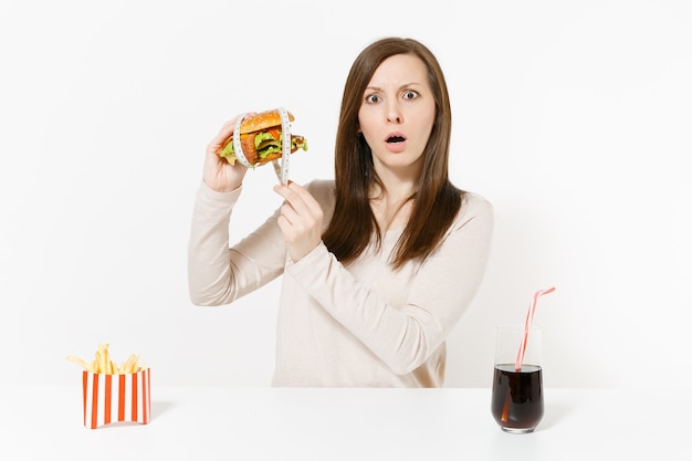 Mulher na mesa segura hambúrguer com alfaiate, fita métrica, batatas fritas, coca-cola em frasco de vidro, isolado no fundo branco. Nutrição adequada ou fast food clássico americano. Área com espaço de cópia.