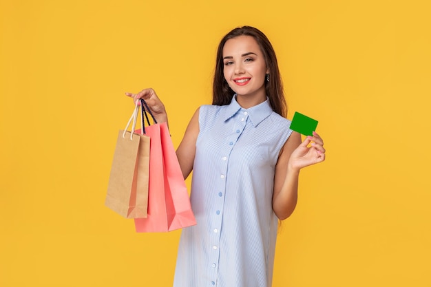 Mulher na moda com 20 anos em um vestido com longos cabelos castanhos sorrindo enquanto segura diferentes pacotes de compras e um cartão nas mãos isolados sobre fundo amarelo