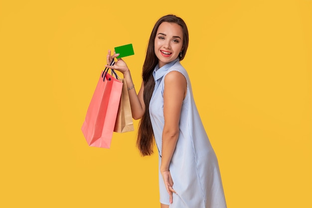 Mulher na moda com 20 anos em um vestido com longos cabelos castanhos sorrindo enquanto segura diferentes pacotes de compras e um cartão nas mãos isolados sobre fundo amarelo