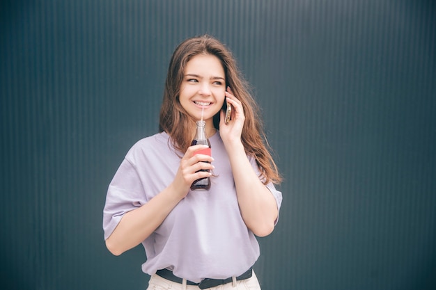 Mulher na moda elegante jovem isolada sobre fundo azul cinza. Menina falando no telefone e bebendo refrigerante de água com gás escuro de garrafa de vidro por canudo plástico.