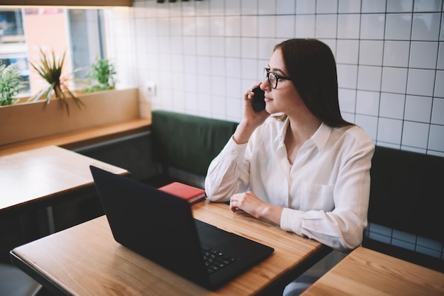 Mulher na moda falando por smartphone com colega e usando laptop enquanto está sentado no espaço de coworking