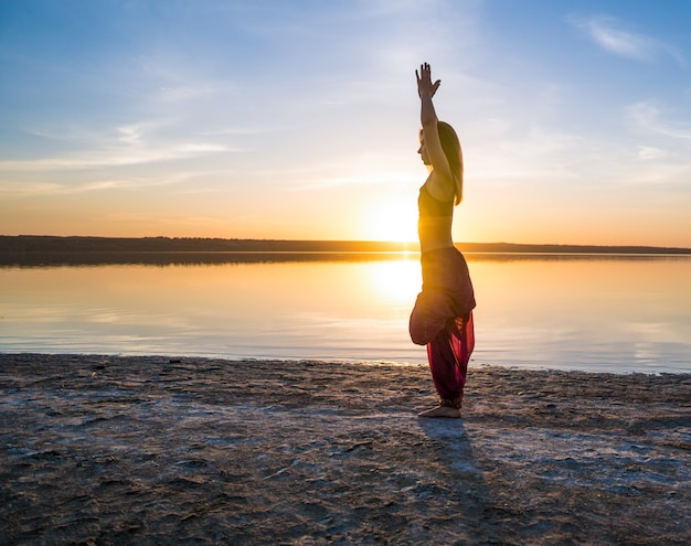 Mulher na praia ao pôr do sol começa a fazer treinamento de ioga asana. Treino de alongamento natural matinal de aquecimento