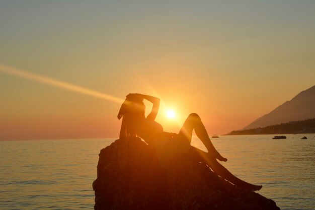 Foto mulher na rocha ao lado do mar contra o céu durante o pôr do sol