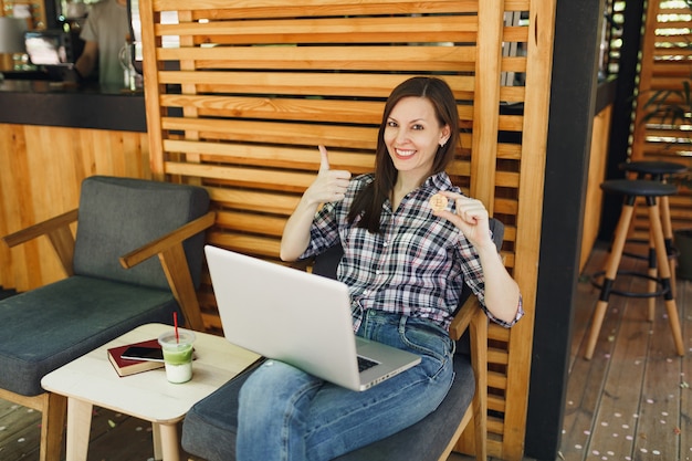 Mulher na rua ao ar livre, café, café, sentada com o computador laptop pc, segura bitcoin, moeda de metal dourada