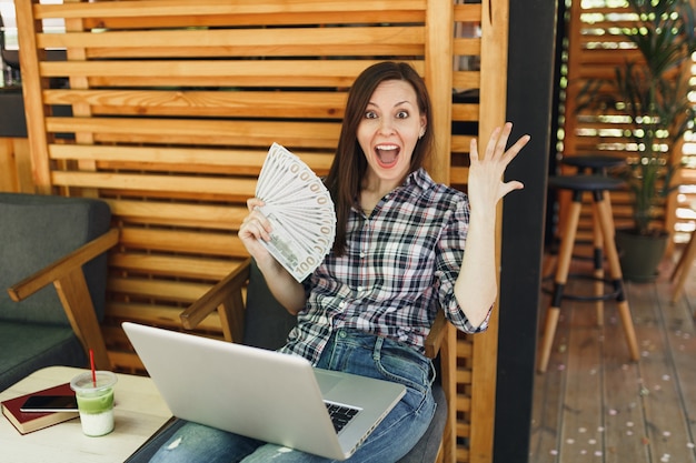 Mulher na rua ao ar livre, café, café, sentada com um computador laptop moderno, segurando um monte de notas de dólares, dinheiro em espécie