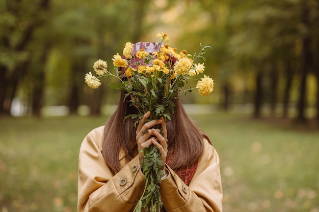 Mulher na trincheira se escondendo atrás do buquê de flores amarelas e cobrindo o rosto em pé no parque