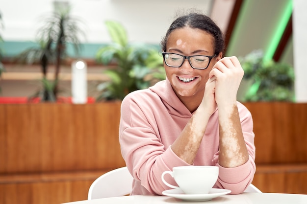 Mulher negra afro-americana com vitiligo pigmentação problema de pele coberta com capuz rosa óculos com capuz sentado na mesa bebida interna chá sorrindo Feliz pessoa positiva esperando no restaurante