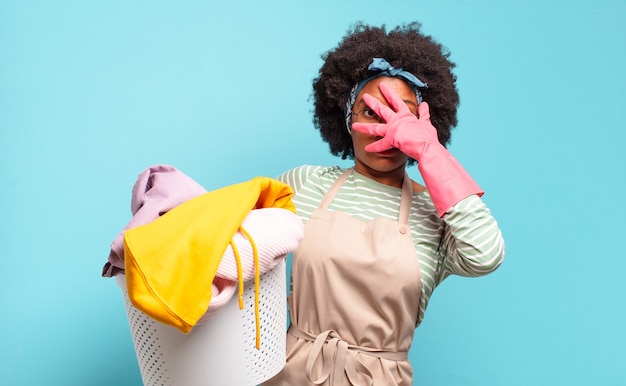 Mulher negra afro parecendo chocada, assustada ou apavorada, cobrindo o rosto com a mão e espiando por entre os dedos. conceito de limpeza. conceito doméstico