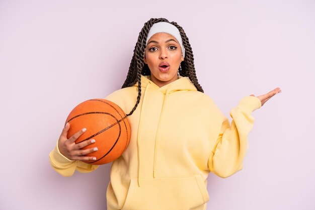 Mulher negra afro parecendo surpresa e chocada com o queixo caído segurando um conceito de basquete de objeto