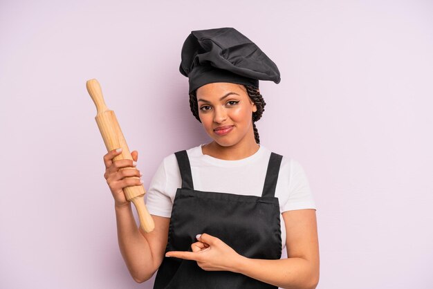 Mulher negra afro sorrindo alegremente, sentindo-se feliz e apontando para o lado. chef com um pino de rolo