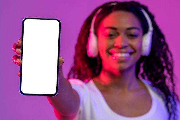 Mulher negra alegre em fones de ouvido sem fio segurando smartphone em branco com tela branca