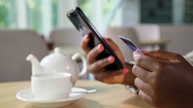 Mulher negra alegre em jaqueta elegante paga online com cartão de crédito e celular, sentada à mesa com bule e xícara em uma cafeteria aconchegante