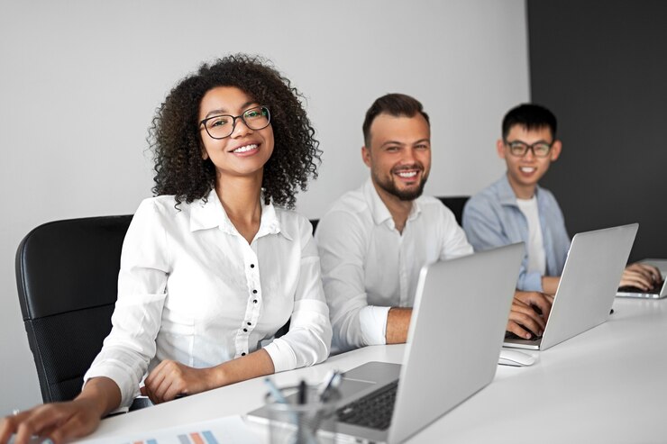Foto mulher negra alegre sorrindo e olhando para a câmera enquanto trabalhava com colegas multirraciais do sexo masculino com laptops no local de trabalho contemporâneo