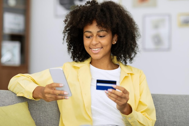 Mulher negra alegre usando smartphone fazendo compras on-line, segurando cartão de crédito, fazendo o pagamento enquanto está sentado