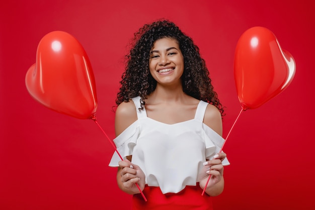 Foto mulher negra animada com balões em forma de coração isolados na parede vermelha