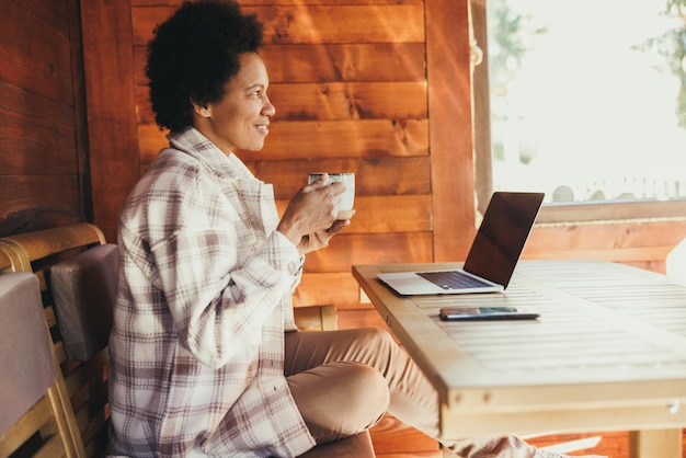 Mulher negra bebendo chá da manhã e usando laptop enquanto passa o tempo de férias em uma aconchegante casa de madeira.