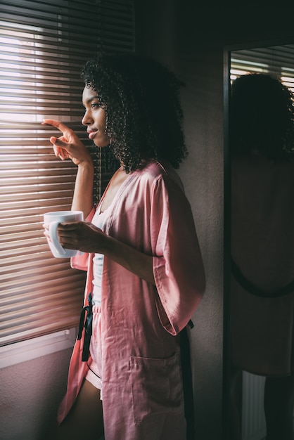 Mulher negra bonita em pé na janela com uma xícara de café