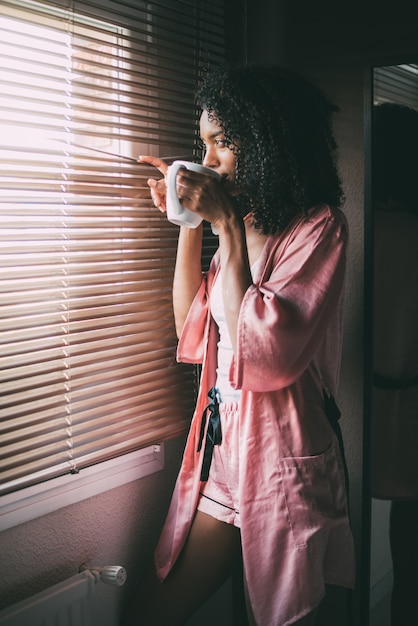 Mulher negra bonita em pé na janela com uma xícara de café