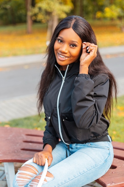 Mulher negra bonita feliz com um sorriso em uma jaqueta casual da moda com jeans, sentada em um banco em um parque de outono com folhagem amarela