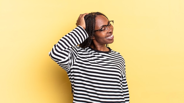 Mulher negra bonita sorrindo alegre e casualmente, levando a mão na cabeça com um olhar positivo, feliz e confiante