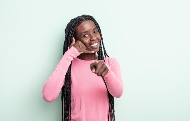 Mulher negra bonita sorrindo alegremente e apontando para a câmera enquanto faz um gesto para ligar para você, falando ao telefone