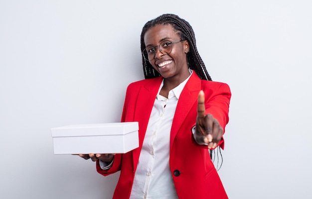 Mulher negra bonita sorrindo com orgulho e confiança fazendo o número um. empresária com uma caixa branca