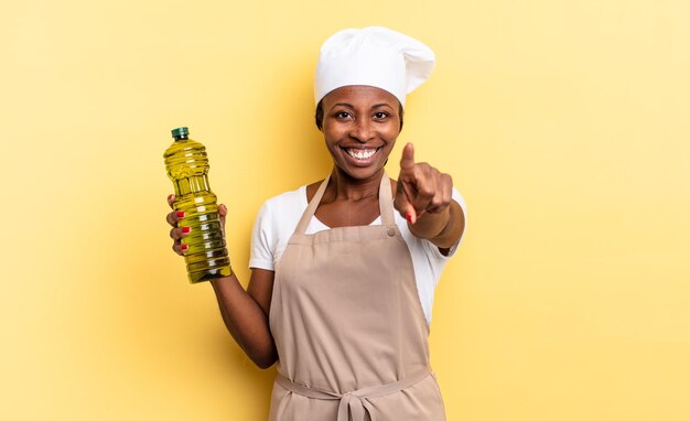 Mulher negra chef afro apontando para a câmera com um sorriso satisfeito, confiante e amigável, escolhendo você. conceito de azeite