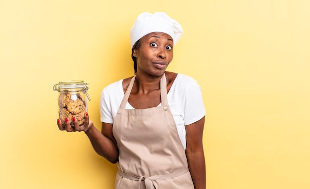 Mulher negra chef afro encolhendo os ombros, sentindo-se confusa e incerta, duvidando com os braços cruzados e olhar perplexo. conceito de biscoitos