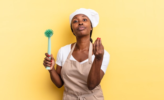 Mulher negra chef afro fazendo capice ou gesto de dinheiro, dizendo para você pagar suas dívidas !. conceito de limpeza de pratos