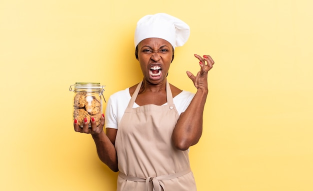 Mulher negra chef afro gritando com as mãos no ar, sentindo-se furiosa, frustrada, estressada e chateada. conceito de biscoitos