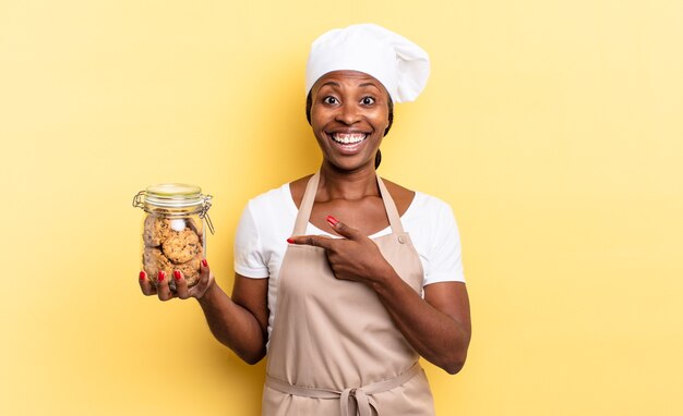 Mulher negra chef afro parecendo animada e surpresa, apontando para o lado e para cima para copiar o espaço. conceito de biscoitos