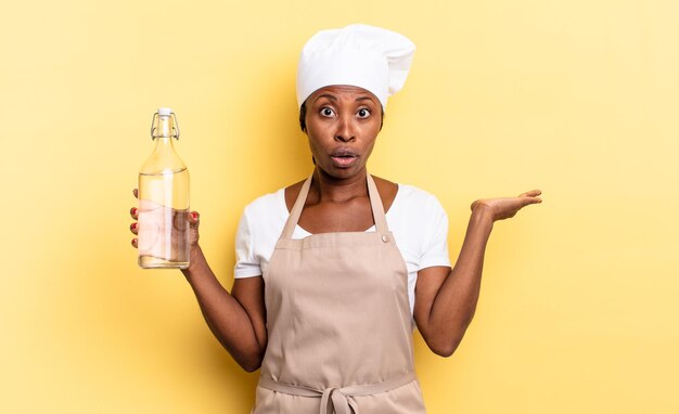 Mulher negra chef afro parecendo surpresa e chocada, com o queixo caído segurando um objeto com a mão aberta segurando uma garrafa de água