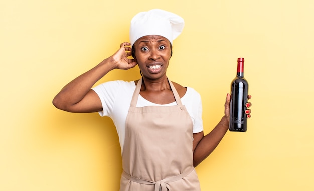 Mulher negra chef afro se sentindo estressada, preocupada, ansiosa ou com medo, com as mãos na cabeça, entrando em pânico por engano. conceito de garrafa de vinho