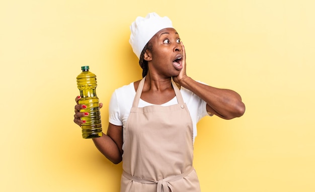 Mulher negra chef afro se sentindo feliz, animada e surpresa, olhando para o lado com as duas mãos no rosto. conceito de azeite