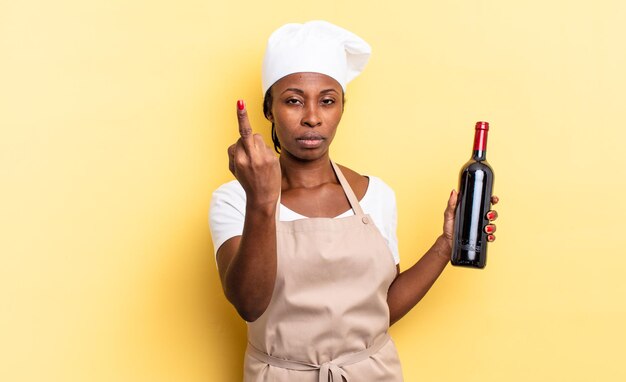 Mulher negra chef afro se sentindo irritada, irritada, rebelde e agressiva, sacudindo o dedo do meio, lutando contra ela. conceito de garrafa de vinho