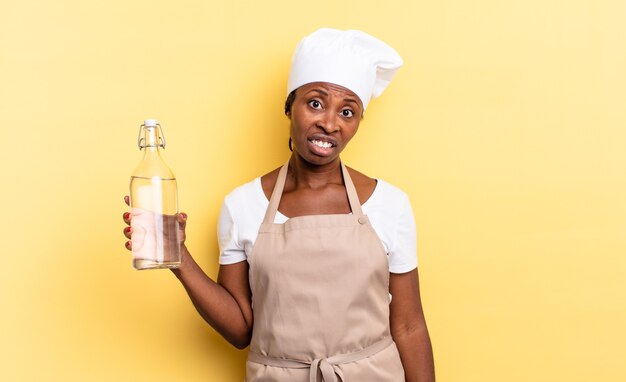 Mulher negra chef afro se sentindo perplexa e confusa, com uma expressão estúpida e atordoada olhando para algo inesperado segurando uma garrafa de água