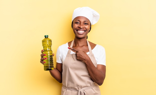 Mulher negra chef afro sorrindo alegremente, sentindo-se feliz e apontando para a lateral e para cima, mostrando o objeto no espaço da cópia. conceito de azeite